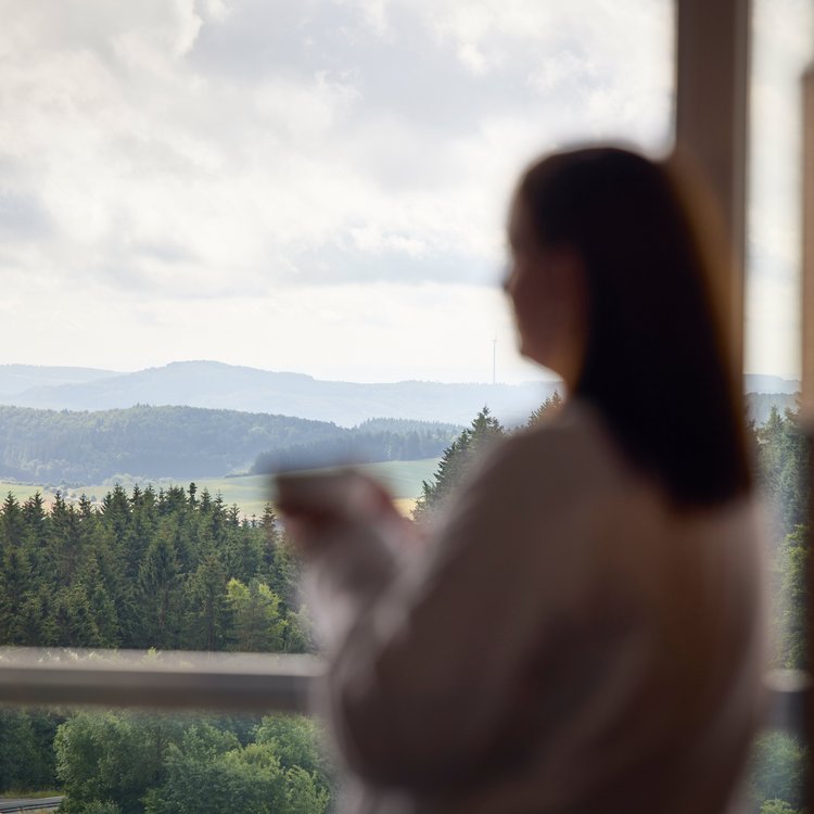 Frau genießt den Ausblick auf die Eifel vom Zimmer im Nürburgring Hotel mit einer Tasse in der Hand
