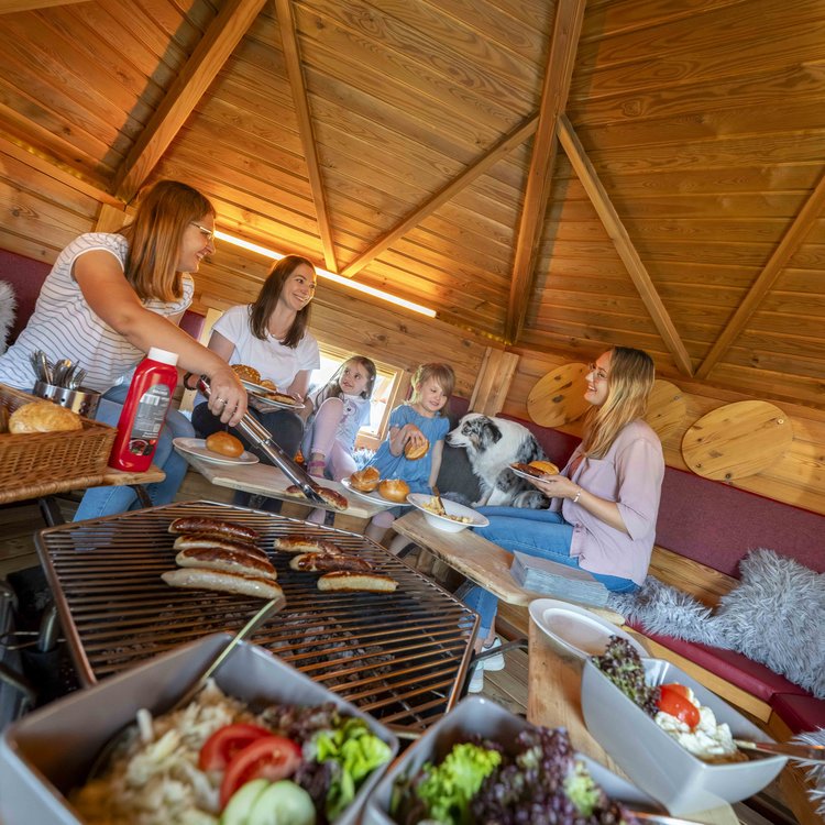 Familie grillt gemeinsam in einer Holzhütte im Nürburgring Ferienpark, gemütliche Atmosphäre mit Kindern und Hund