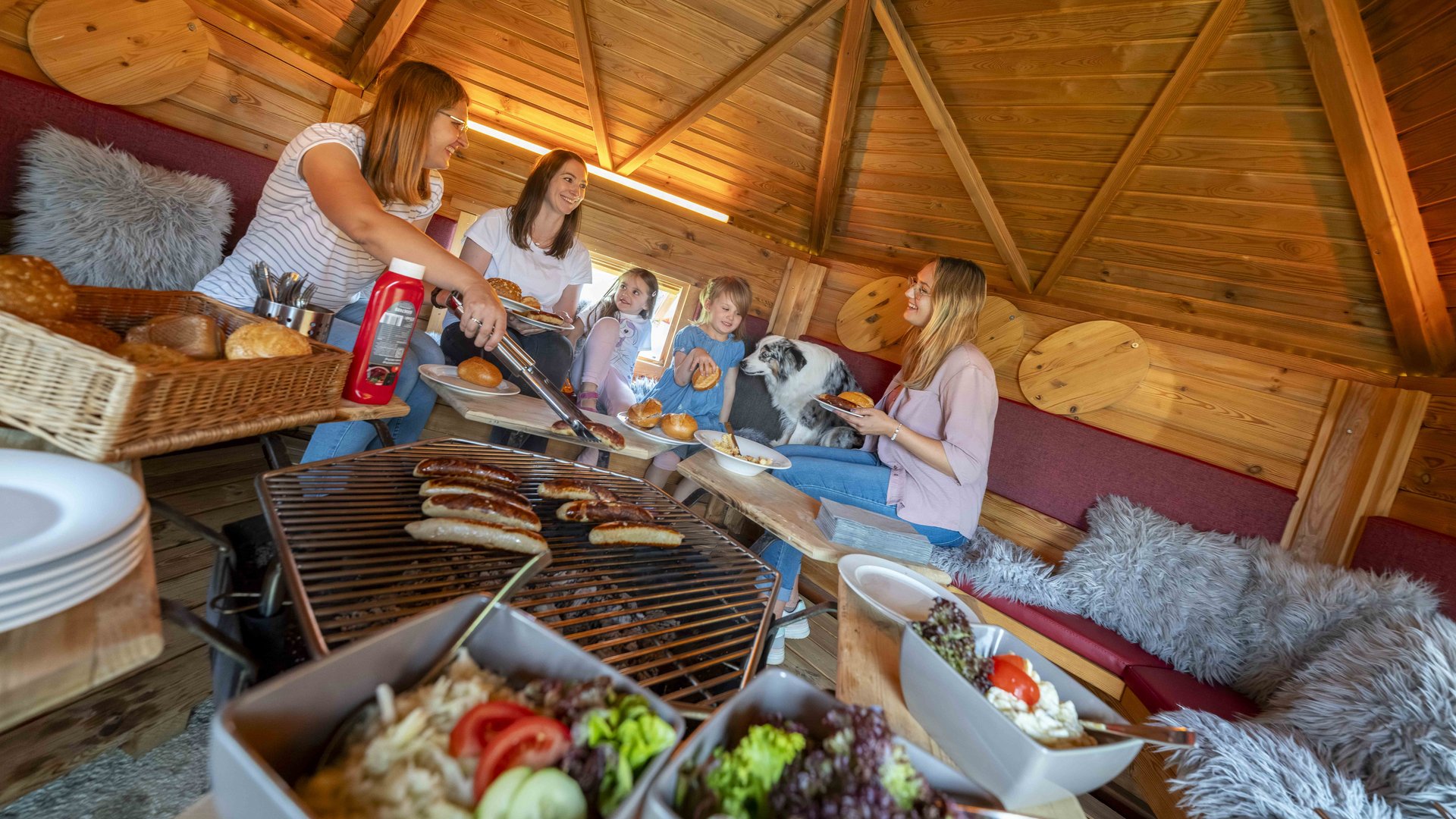 Familie grillt gemeinsam in einer Holzhütte im Nürburgring Ferienpark, gemütliche Atmosphäre mit Kindern und Hund