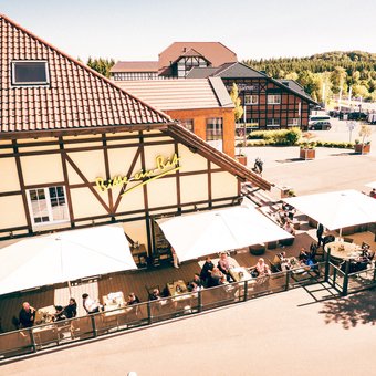 Außenansicht des Bitburger Gasthauses am Nürburgring mit sonniger Terrasse und Gästen unter weißen Schirmen.