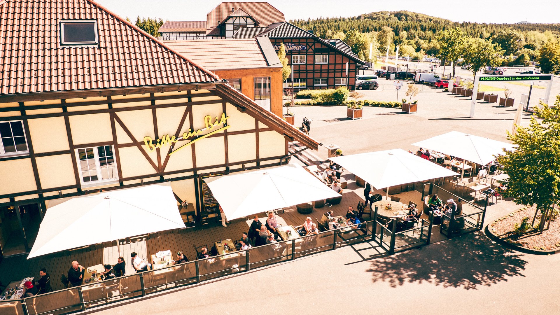 Außenansicht des Bitburger Gasthauses am Nürburgring mit sonniger Terrasse und Gästen unter weißen Schirmen.