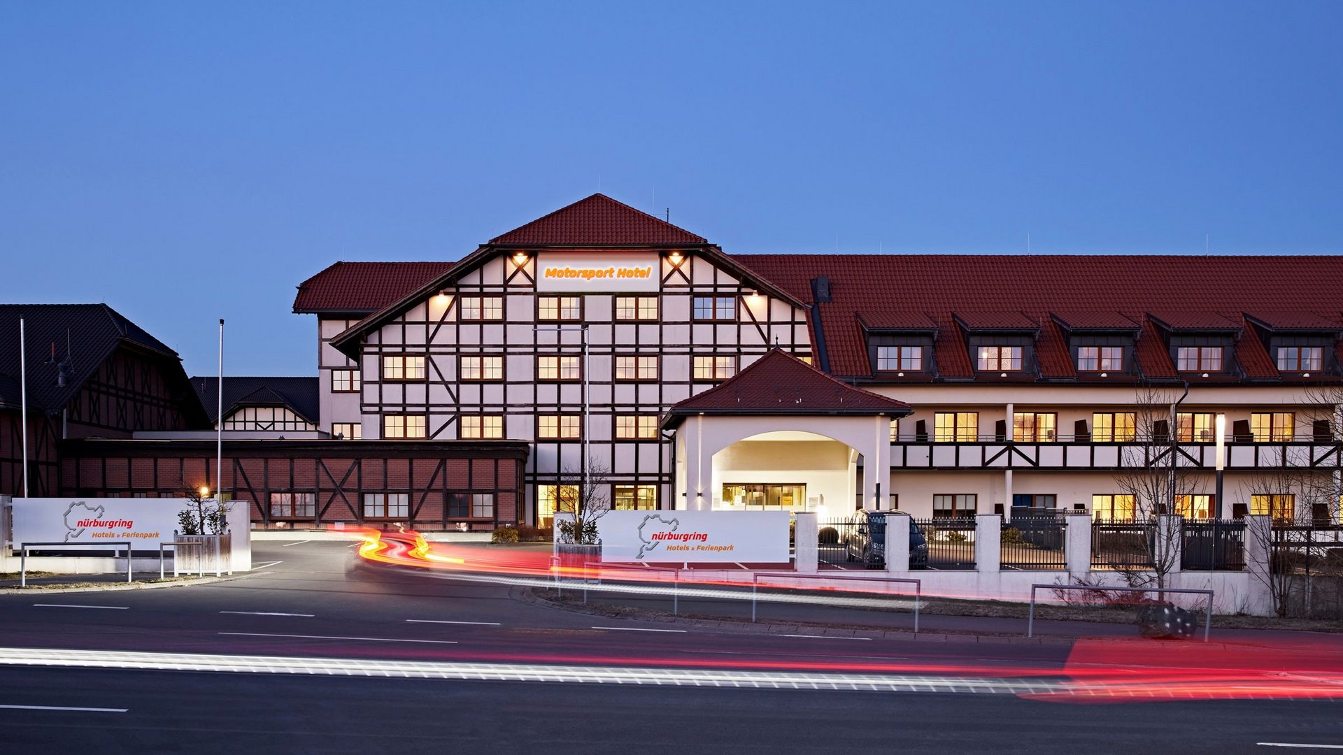 Außenansicht des Nürburgring Motorsport Hotels in der Abenddämmerung mit beleuchteter Fassade und Parkplatz.