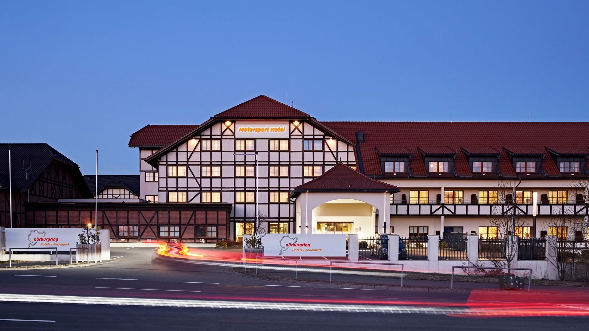 Außenansicht des Nürburgring Motorsport Hotels in der Abenddämmerung mit beleuchteter Fassade und Parkplatz.
