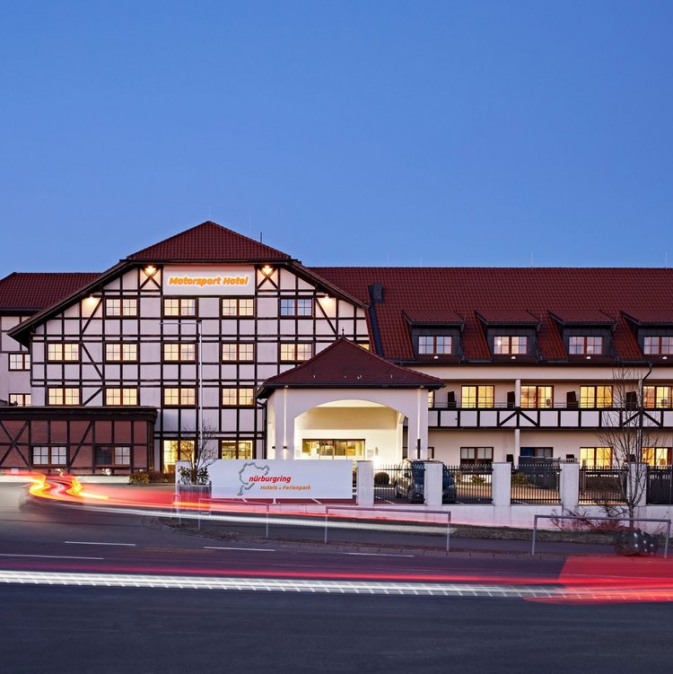 Außenansicht des Nürburgring Motorsport Hotels in der Abenddämmerung mit beleuchteter Fassade und Parkplatz.