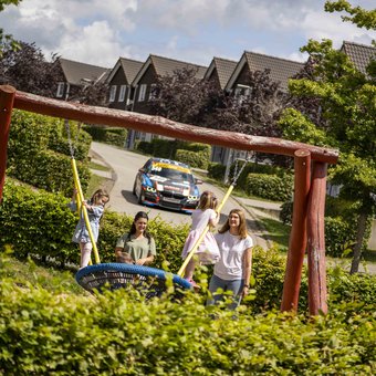 Familienurlaub im Nürburgring Ferienpark – Kinder spielen auf der Schaukel, Rennwagen im Hintergrund